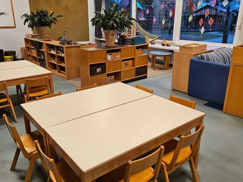 Ein farbenfroher Kindergartenraum mit Tischen, Stühlen, Regalen und großen Fenstern, die geschmückt sind.