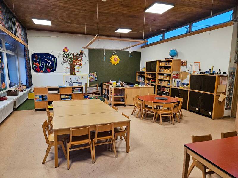 Ein heller Kindergartenraum mit Holzmöbeln, mehreren Tischen und Stühlen, einem Teppich in der Mitte und Regalen voller Spielsachen und Büchern.