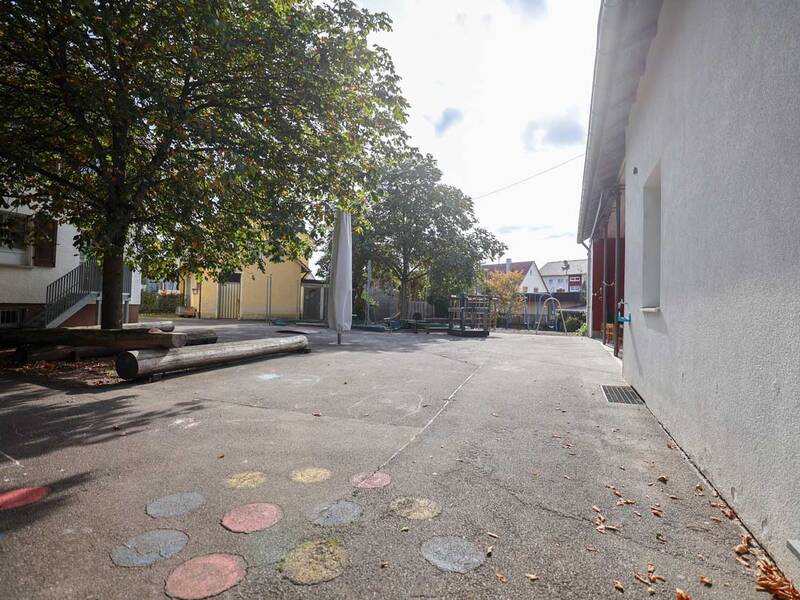Ein leerer Schulhof mit bunten Kreidemalereien, einem großen Baum links und Gebäuden im Hintergrund.