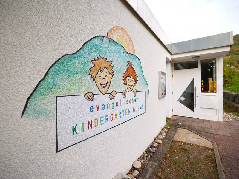 Ein Wandbild mit zwei lächelnden Kindern und dem Schriftzug „Evangelischer Kindergarten Glems“ ist auf der Außenwand eines Gebäudes nahe dem Eingang zu sehen.
