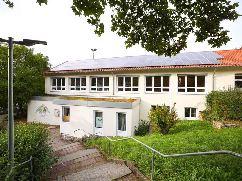 Ein zweistöckiges Gebäude mit weißen Wänden und großen Fenstern, auf dessen orangefarbenem Ziegeldach Solarpaneele installiert sind, umgeben von grüner Landschaft.