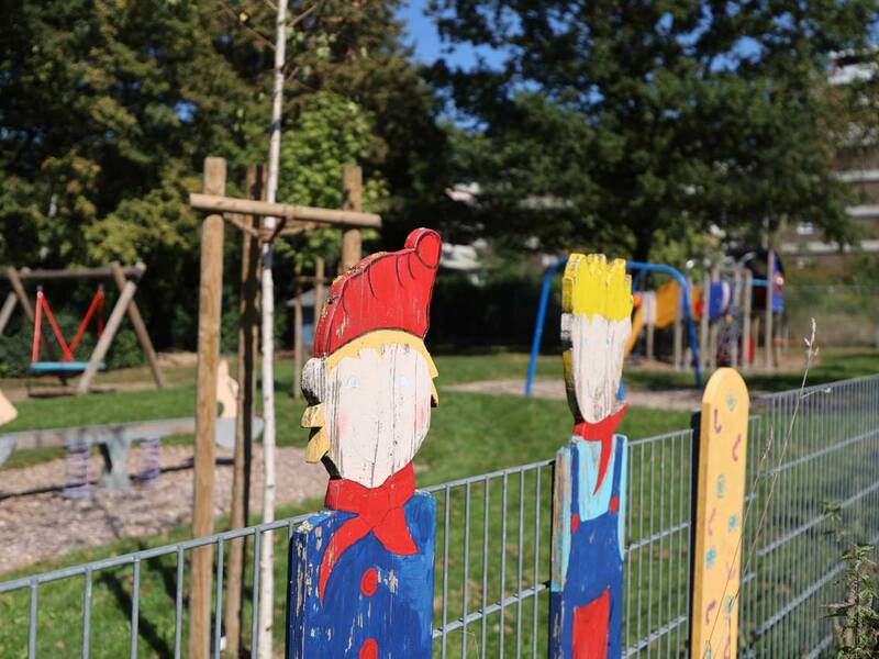 Zwei bunt bemalte Holzfiguren sind auf einem Zaun vor einem Spielplatz mit Schaukeln und Klettergerüsten zu sehen.