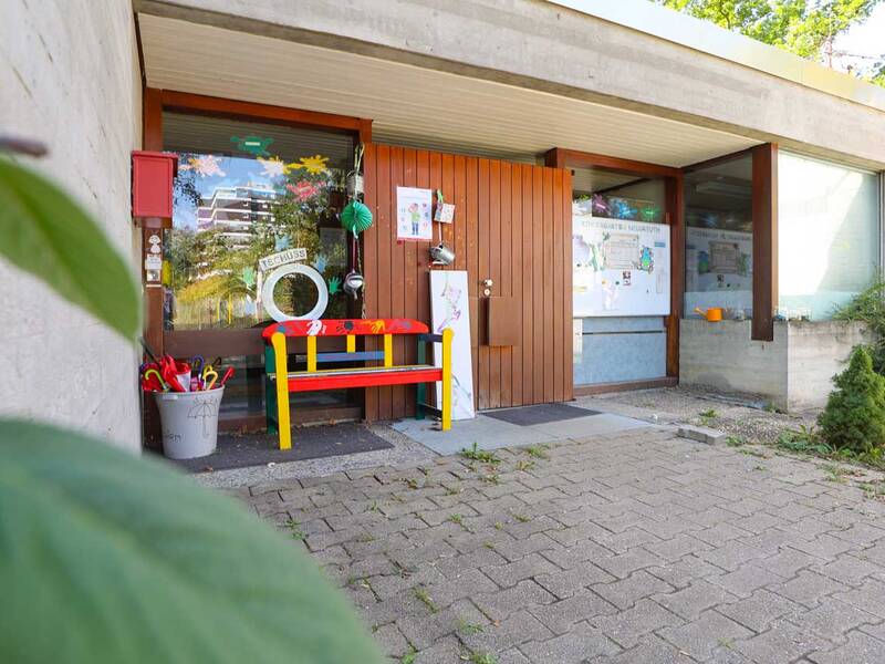 Eingangsbereich eines Gebäudes mit großer Holztür, bunter Bank, Dekorationen und Pflanzendetails im Vordergrund.
