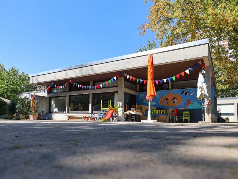 Ein flaches Gebäude mit einem bunt bemalten Wandbild und bunten Wimpelketten, daneben Kinderstühle und ein Spielplatzrutsche, umgeben von Bäumen und Sonnenschirmen.