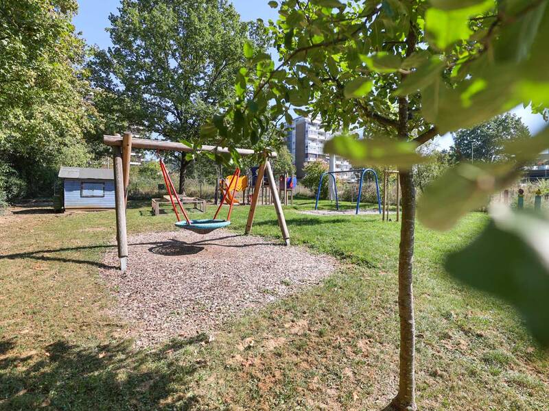 Ein Spielplatz mit einer Nestschaukel, einem kleinen blauen Häuschen und weiteren Spielgeräten befindet sich auf einer grünen Wiese mit Bäumen im Hintergrund.