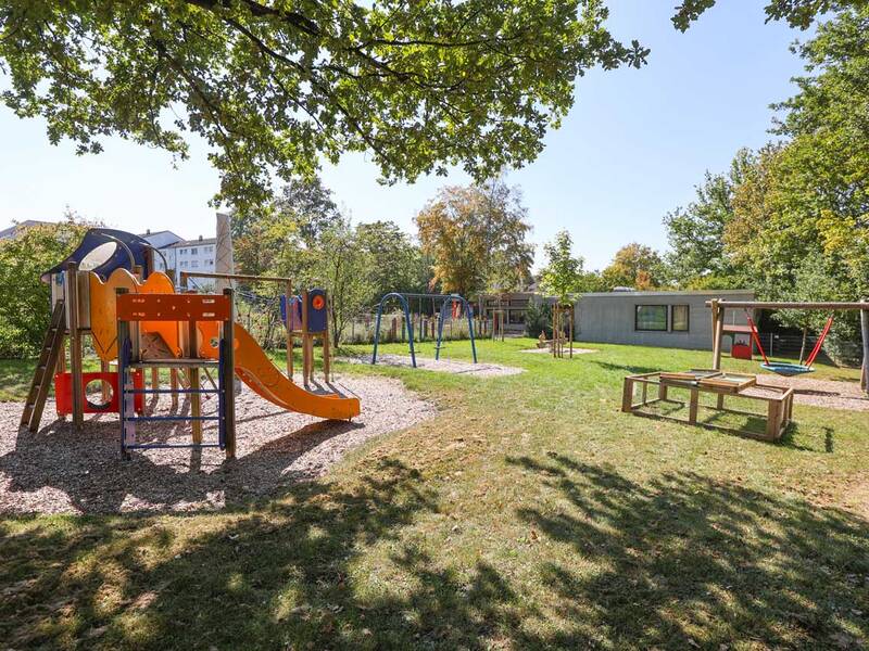 Ein Spielplatz mit orangener Rutsche und Klettergeräten steht auf einer Wiese, umgeben von Bäumen und einem Gebäude im Hintergrund.