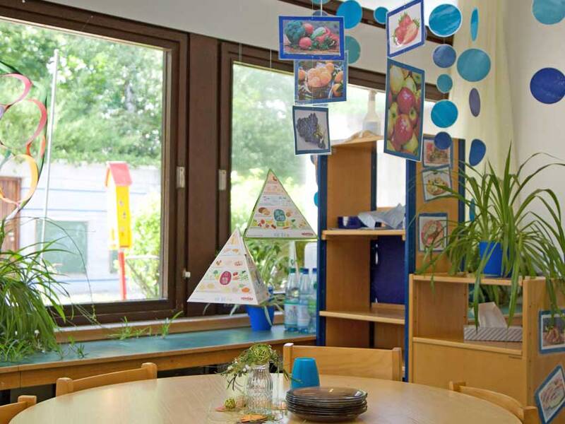 Ein heller Raum mit einem runden Holztisch, umgeben von kleinen Holzstühlen, an dem bunte Bilder von Lebensmitteln von der Decke hängen und Pflanzen auf der Fensterbank stehen.