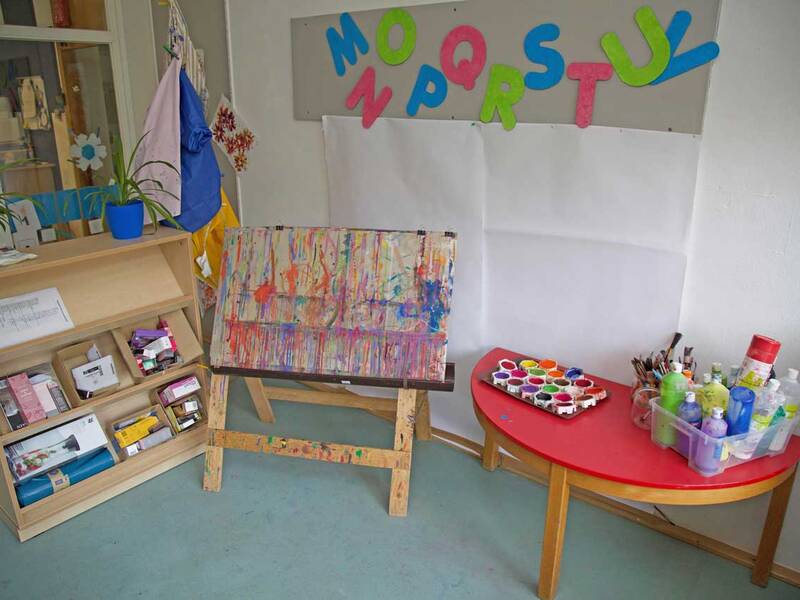Ein Raum mit einem Holzregal voller Bastelmaterialien, einer Staffelei mit einer bemalten Leinwand und einem Tisch mit verschiedenen Malutensilien und Farben.