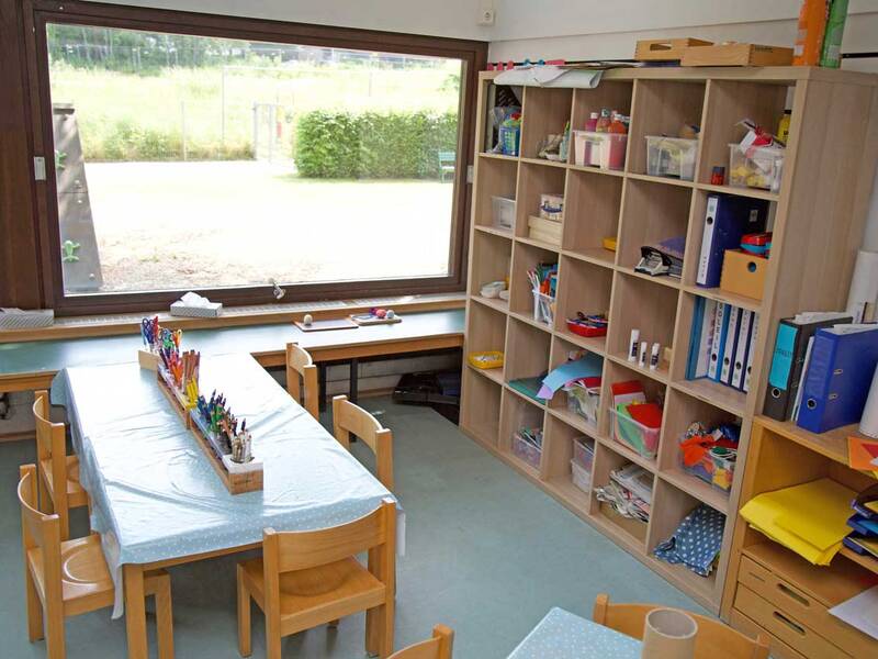 Ein heller Raum mit großen Fenstern enthält einen Tisch mit Kindersitzen und Regalen voller Bastelmaterialien.