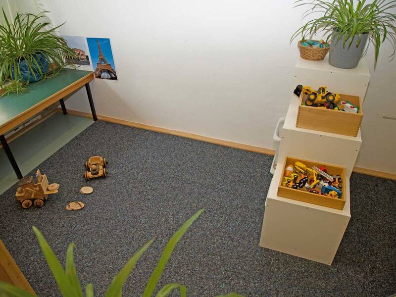 Ein Spielzimmer mit grauem Teppichboden, auf dem ein Holzwagen und Bausteine liegen, sowie kleine Regale mit verschiedenen Spielzeugen und Pflanzen stehen.