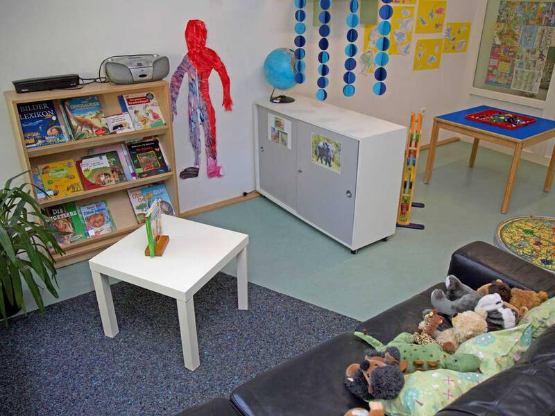 Ein gemütlicher Kinderbereich mit einer Couch voller Stofftiere, einem Bücherregal, einem kleinen Tisch und bunten Dekorationen an Wänden und Decke.