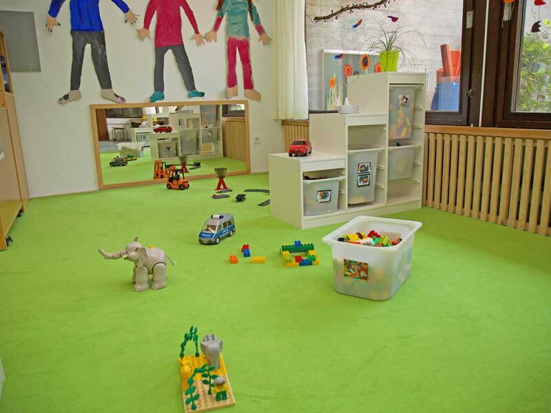 Ein farbenfroher Kindergartenraum mit grünem Teppichboden ist mit verschiedenen Spielzeugen, wie einem Elefanten, einem Polizeiauto und einer Box mit Bausteinen, sowie bunten Wanddekorationen gestaltet.