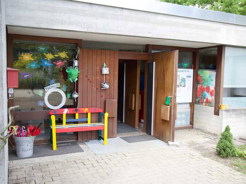 Der Eingang eines Kindergartens ist mit bunten Bildern und einer farbenfrohen Bank geschmückt, und eine Tür steht einladend offen.