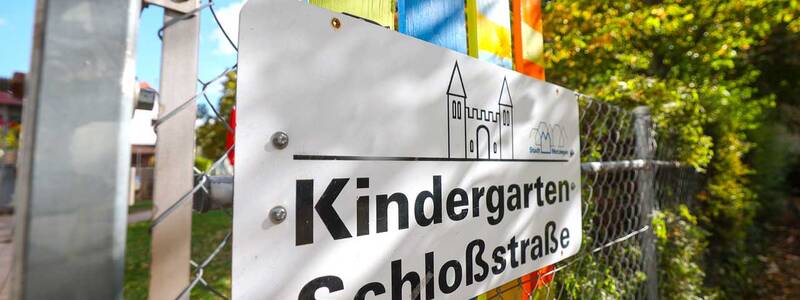 Nahaufnahme eines Schilds am Zaun. Darauf steht „Kindergarten Schlossstraße“, daneben eine einfache Zeichnung einer Burg. Hinter dem Schild sind bunte Holzlatten in Gelb, Blau, Grün und Rot zu sehen.