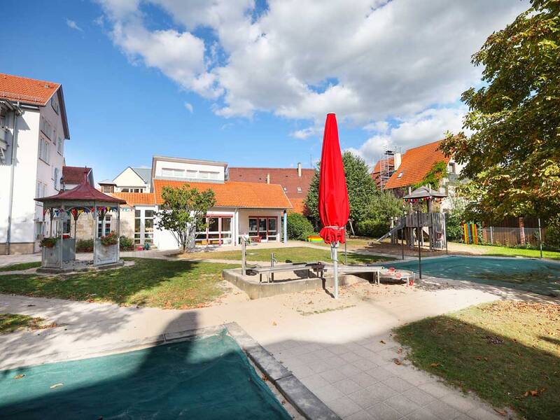Blick über den Garten mit verschiedenen Spielbereichen – ein Pavillon, ein Wasser-Matsch-Bereich mit geschlossenem Sonnenschirm und eine große Holzspielanlage. Dazwischen Wiese, Bäume und helle Häuser mit roten Dächern.