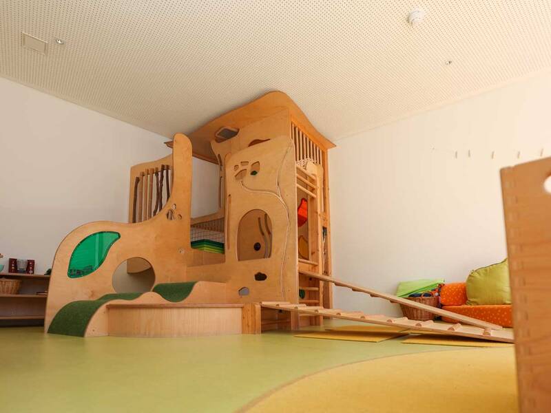 Ein Indoor-Spielbereich mit einer hölzernen Kletterstruktur und einer Rutsche ist in einem hellen Raum aufgebaut.