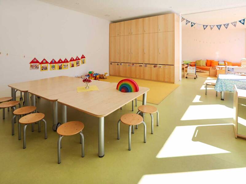 Ein heller und geräumiger Kindergartenraum mit Holztischen, kleinen Stühlen, buntem Spielzeug und einem großen Schrank an der Wand.