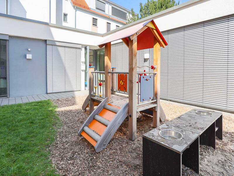 Ein moderner Spielplatz mit einem kleinen Holzspielhaus und Rutschen befindet sich neben einem Gebäude mit geschlossenen Jalousien.