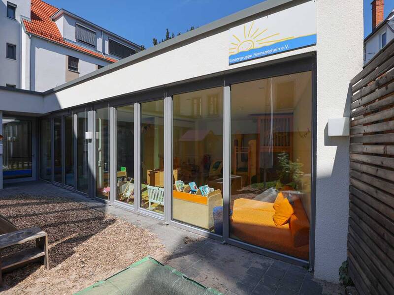 Das Bild zeigt den Eingangsbereich einer Kindertagesstätte mit großen Fenstern, durch die ein Innenraum mit buntem Kinderzimmermobiliar zu sehen ist.