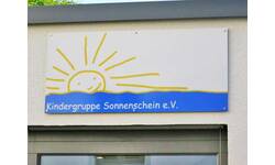 Ein Schild mit der Aufschrift "Kindergruppe Sonnenschein e.V." zeigt eine Zeichnung einer lachenden Sonne, die über einer blauen Welle erscheint.