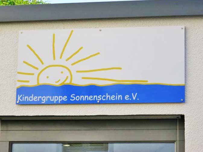 Ein Schild mit der Aufschrift "Kindergruppe Sonnenschein e.V." zeigt eine Zeichnung einer lachenden Sonne, die über einer blauen Welle erscheint.