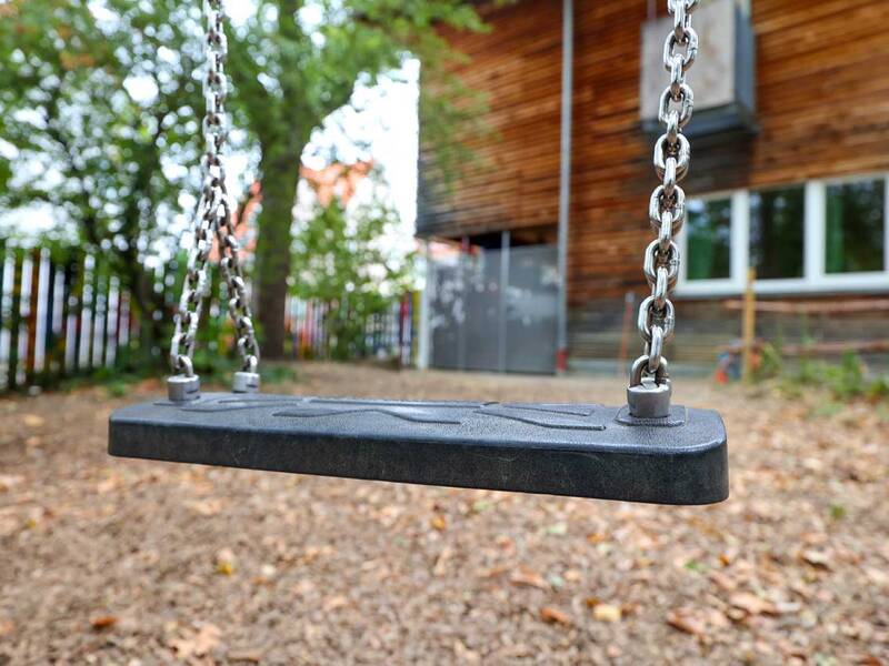 Nahaufnahme einer schwarzen Schaukel mit Metallketten, im Hintergrund Bäume und ein Holzhaus.