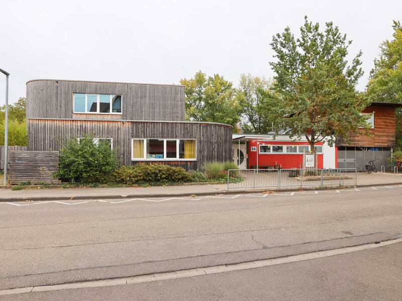 Modernes, teils rundes Holzgebäude mit roten und weißen Akzenten – das Kinderhaus von außen.