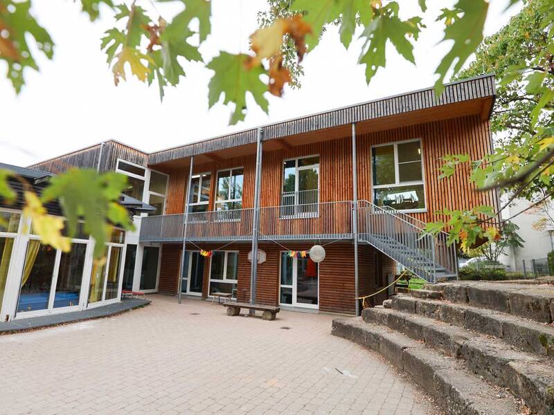 Kindergarten-Gebäude: Ein modernes Holzgebäude mit großen Fenstern und Balkon, davor ein gepflasterter Hof mit Sitzstufen.
