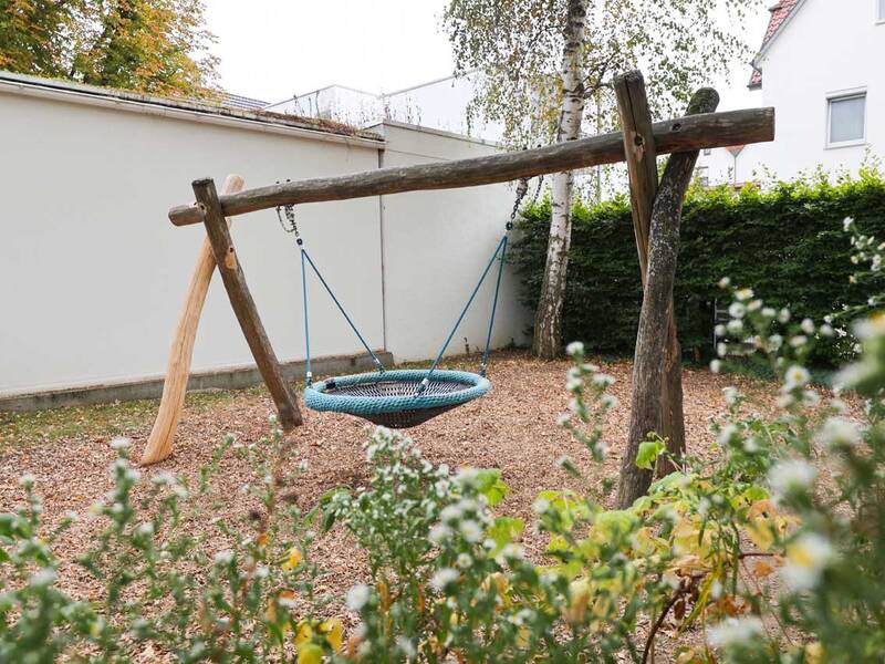 Eine blaue Nestschaukel hängt zwischen zwei rustikalen Holzbalken, umgeben von Bäumen und Hecken.