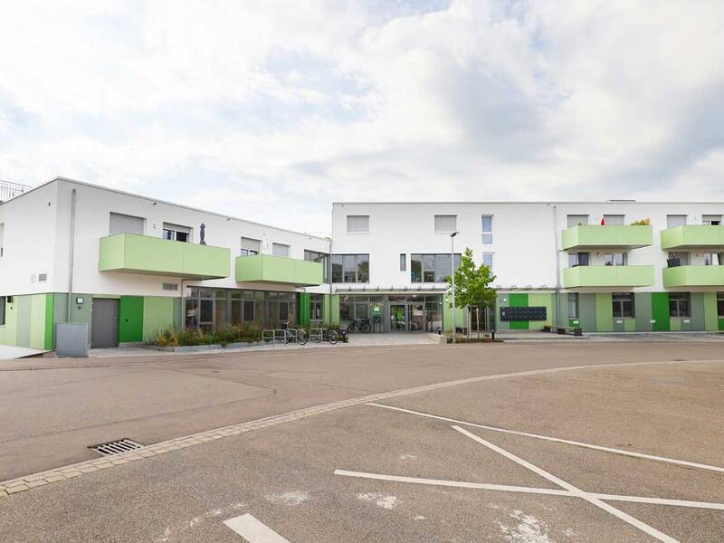 Ein modernes zweistöckiges Gebäude mit grünen Balkonen und Fassadenakzenten steht auf einem großen, leeren Vorplatz unter einem bewölkten Himmel.