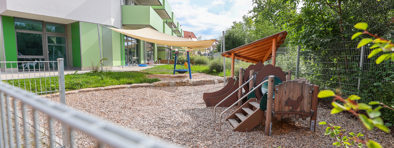 Ein moderner Spielplatz mit Rutschen und Klettermöglichkeiten befindet sich neben einem Gebäude mit grünen Balkonen und Sonnensegeln.