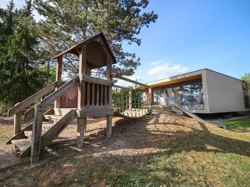 Ein Spielplatz mit Holzklettergerüst und Rutsche steht im Vordergrund, während ein modernes Gebäude mit großen Fenstern im Hintergrund zu sehen ist.