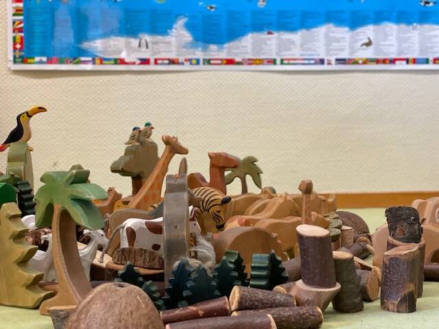 Auf dem Bild sind verschiedene Holztiere, Bäume und Hölzer auf einem Boden verteilt, im Hintergrund hängt eine bunte Weltkarte an der Wand.
