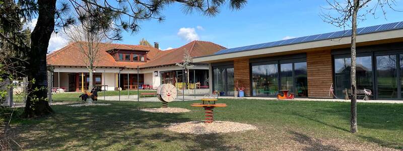 Ein modernes Gebäude mit Holzverkleidung und großen Fenstern steht in einer grünen Umgebung mit Bäumen und einem Spielplatz im Vordergrund.