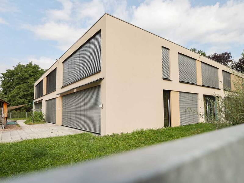 Ein modernes, zweistöckiges Gebäude mit großen Jalousien vor den Fenstern steht neben einem kleinen Spielhaus und einer Grünfläche.