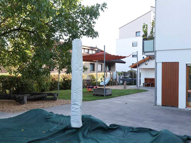 Im Bild ist ein Außenbereich mit gepflastertem Boden, Grasecken, spielenden Strukturen wie einer Rutsche und schattenspendenden Stoffdächern zu sehen, umgeben von modernen Gebäuden und Bäumen.