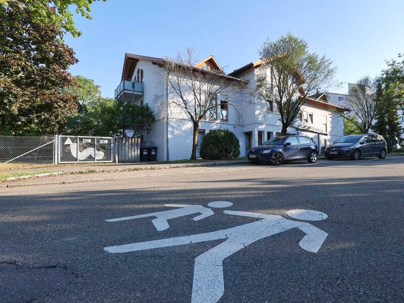 Ein weißes Gebäude mit mehreren Autos davor, Bäume umgeben das Gebäude, und ein Fußgänger-Symbol ist auf die Straße gemalt.