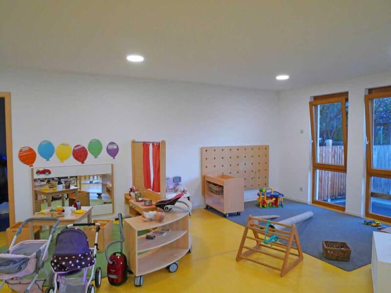 Ein heller Raum mit gelbem Boden und großen Fenstern ist mit verschiedenen Kinderspielzeugen und Möbeln ausgestattet.