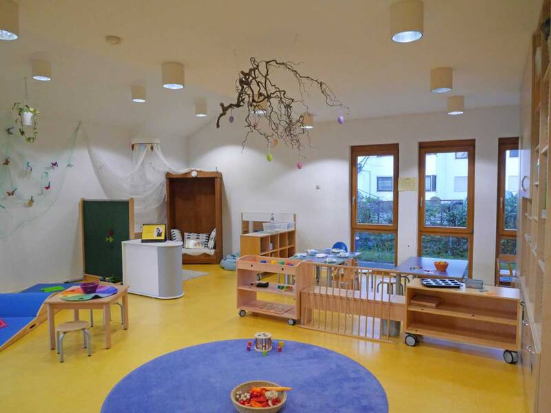 Ein heller Kindergartenraum mit gelbem Boden, blauen Teppichen, kindgerechten Möbeln, Spielsachen und Dekorationen wie hängenden Zweigen und Schmetterlingen.