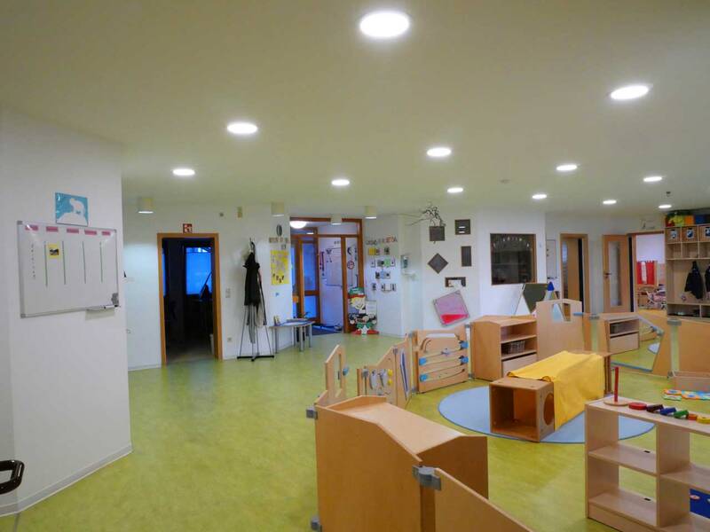 Ein heller Kindergartenraum mit Spielbereichen, Regalen und Kindermöbeln auf einem grünen Boden.