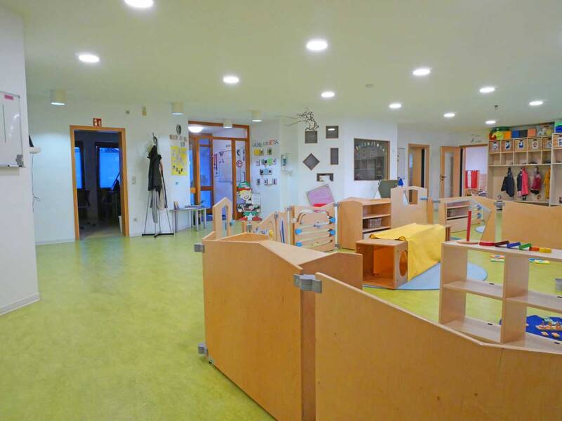 Ein heller und farbenfroher Kindergartenraum mit Spielbereich, Holzspielzeug und bunten Mänteln an der Garderobe.