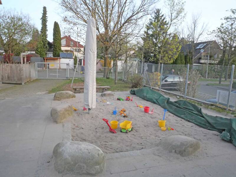 Ein Sandkasten auf einem Spielplatz mit zahlreichen bunten Sandspielzeugen wie Eimern und Schaufeln, umgeben von Bäumen und einem Zaun.