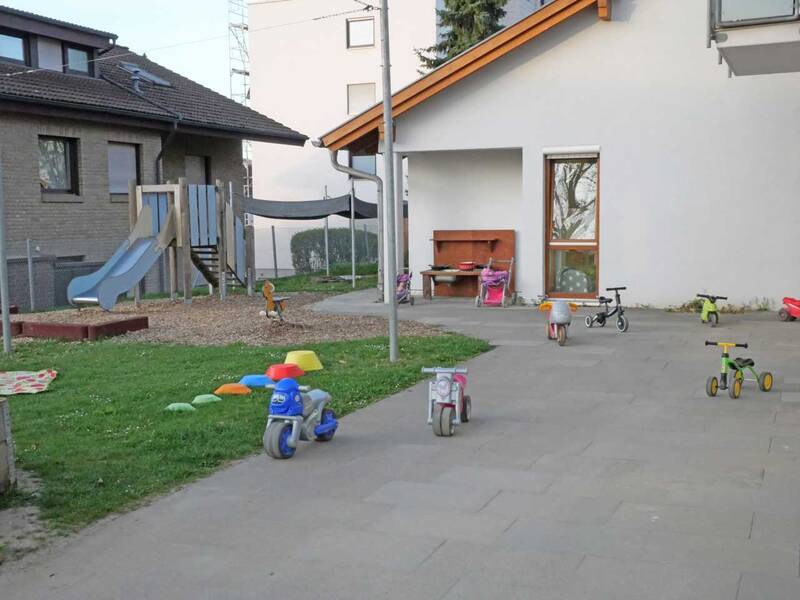 Ein Spielplatz vor einem Haus mit Rutschen, bunten Spielfahrzeugen und Tricycles auf einer gepflasterten Fläche.