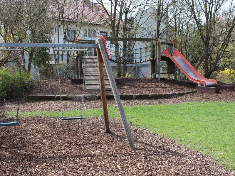 Ein Spielplatz mit Schaukeln, einer Rutsche und einer Kletterstruktur, umgeben von Bäumen und einem Mehrfamilienhaus im Hintergrund.