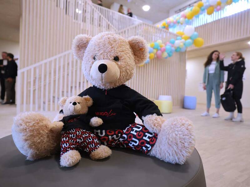 Ein großer Teddybär mit einem kleinen Teddybär auf dem Schoß sitzt vor einer Treppe, während im Hintergrund bunte Luftballons und Menschen zu sehen sind.