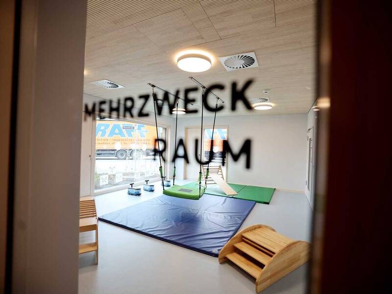 Ein lichtdurchfluteter Mehrzweckraum mit Matten, einer Hängeschaukel und hölzernen Spielgeräten ist durch eine Glastür sichtbar.