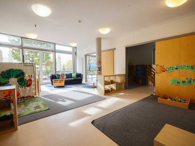 Ein heller, freundlicher Kindergartenraum mit großen Fenstern, bunter Dekoration und Spielzeug, sowie Sitzgelegenheiten.