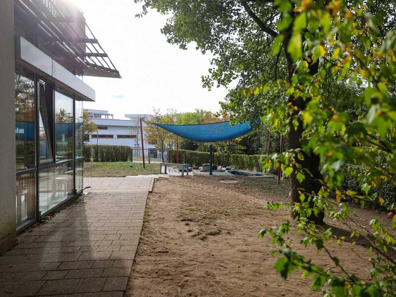 Der Bildausschnitt zeigt links ein Gebäude mit großen Fenstern und rechts einen Sandspielplatz mit einem blauen Sonnensegel, umgeben von Bäumen und Büschen.
