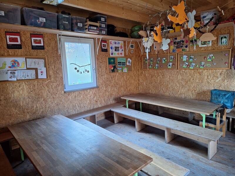 Ein rustikaler Raum mit Holztischen, Bänken und einer Wand aus Korkplatten, die mit Kinderzeichnungen und Bastelarbeiten dekoriert ist.