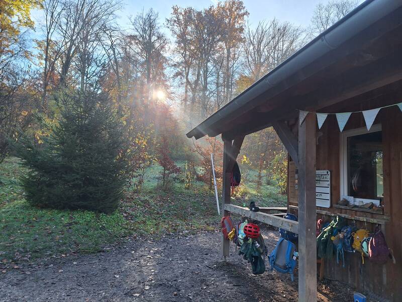 Eine hölzerne Hütte im Wald mit aufgehängten bunten Rucksäcken und einem Sonnenstrahl, der durch die Bäume scheint.